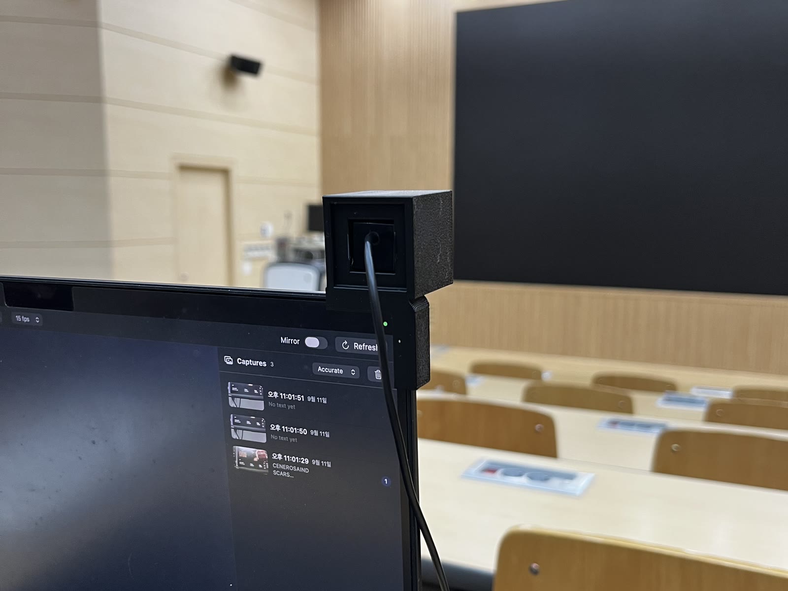 A small black printed cube clipped to the top right corner of a MacBook lid in an empty lecture hall, a USB cable running down from it, the large dark board at the front of the room behind it