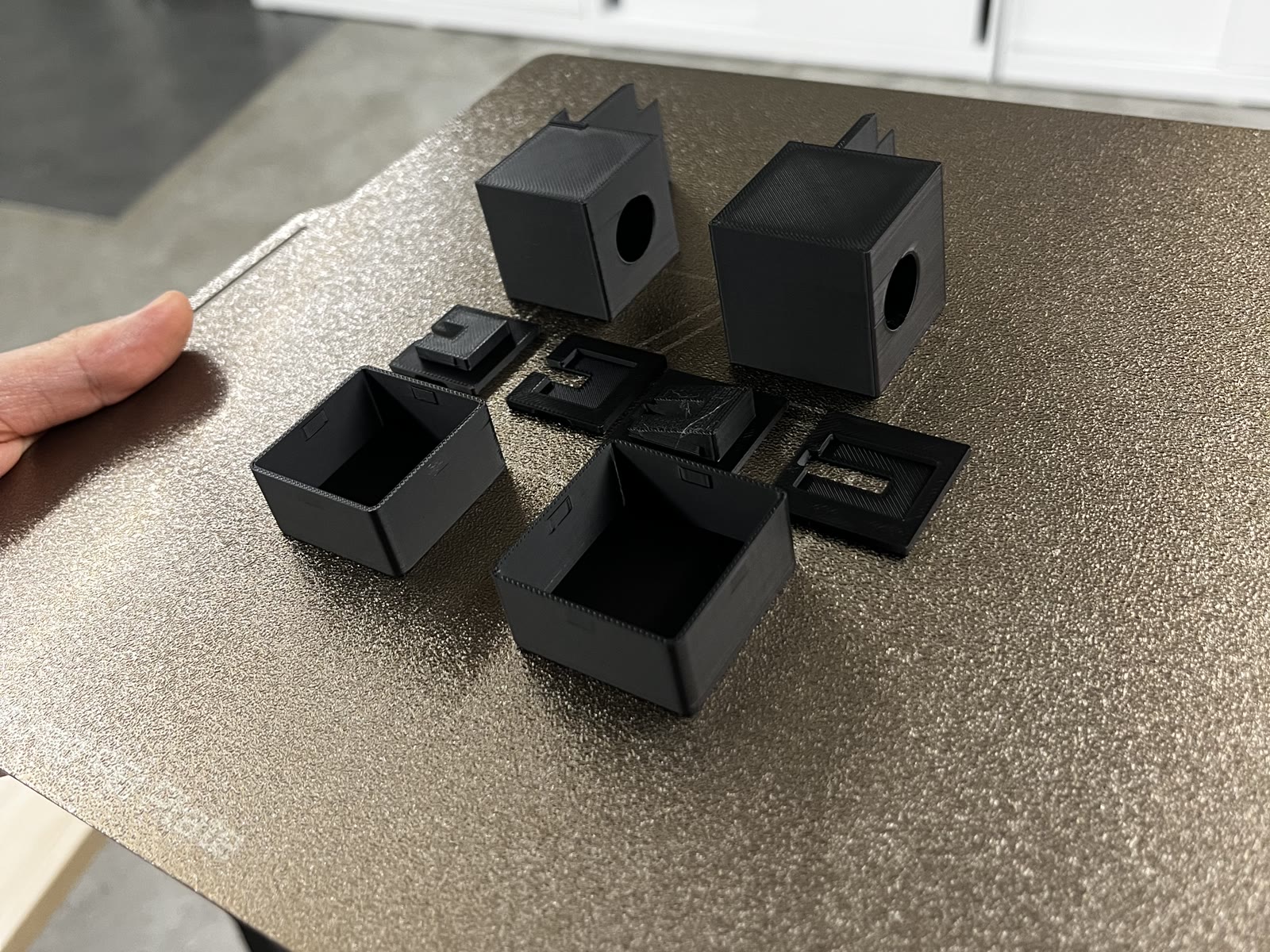Freshly printed black parts on a print bed: two cubes with a lens hole, their hatches, and two open pocket sleeves